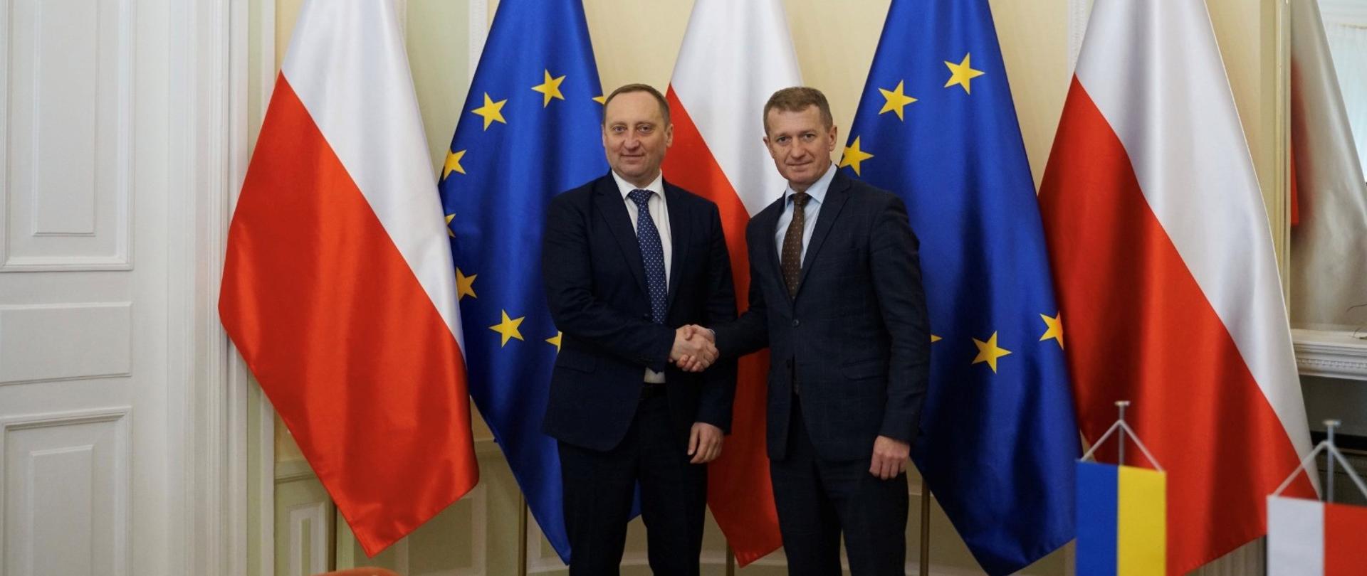Wiceminister Ireneusz Raś oraz wiceminister ds. sportu Ukrainy Andrij Czesnokow podają sobie ręce. W tle flagi Polski i Unii Europejskiej.