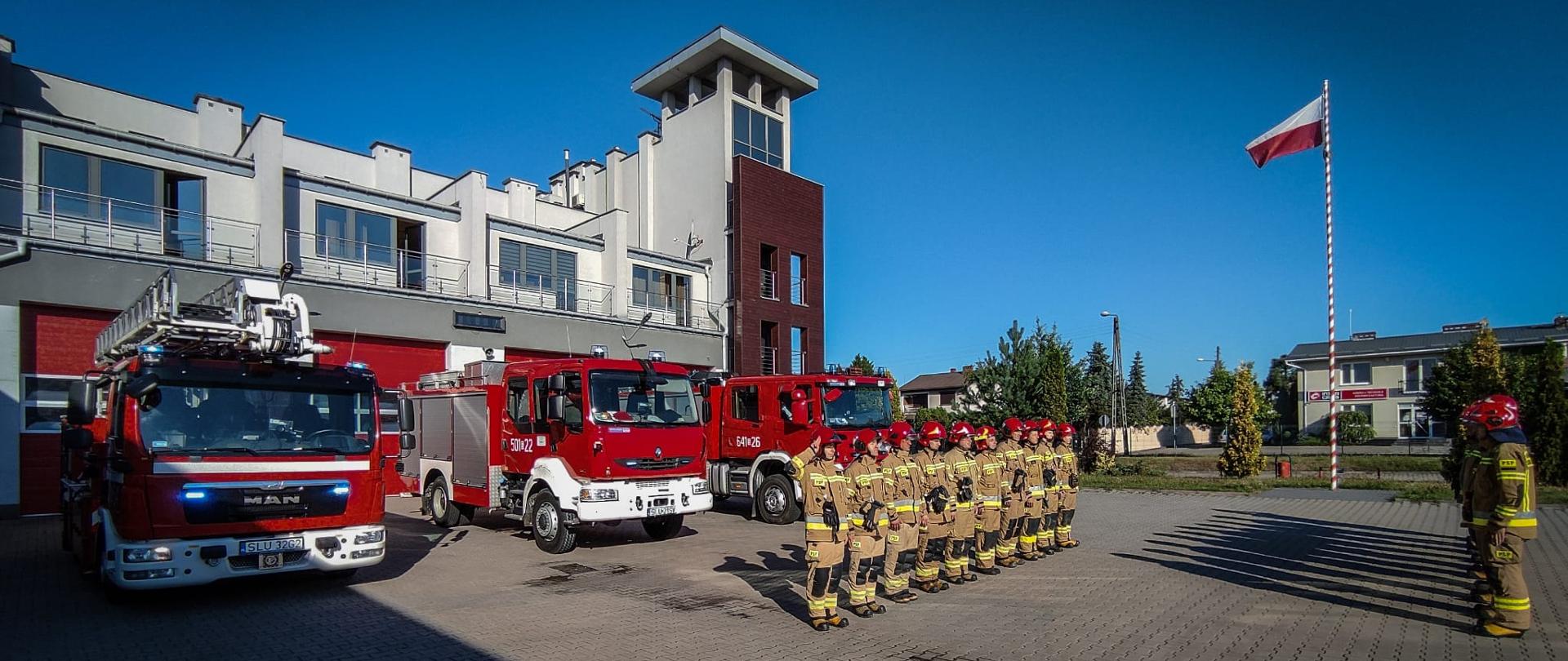 Po prawej stronie zdjęcia strażacy ustawieni w dwóch rzędach po prawej i lewej stronie podczas uroczystej zmiany służby, stojący na baczność, oddają hołd tragicznie zmarłemu strażakowi z Komendy Głównej PSP. Z lewej strony trzy samochody pożarnicze na tle budynku komendy. Po prawej stronie maszt z podniesioną flagą państwową.