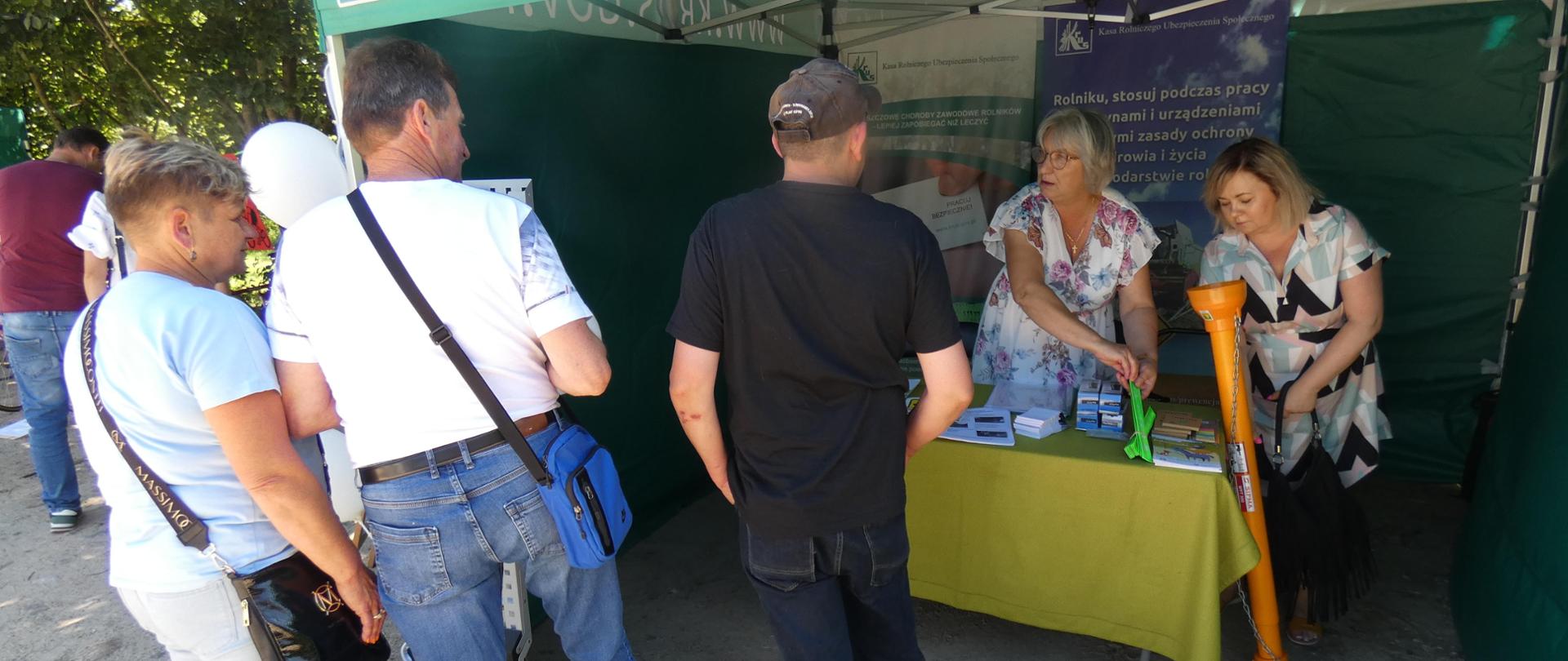 Przed namiotem, pod którym znajduje się stoisko informacyjne Placówki Terenowej KRUS w Siemiatyczach stoją dwaj mężczyźni i jedna kobieta . Wewnątrz namiotu dwie kobiety przekazują odwiedzającym stoisko broszury i ulotki prewencyjne.