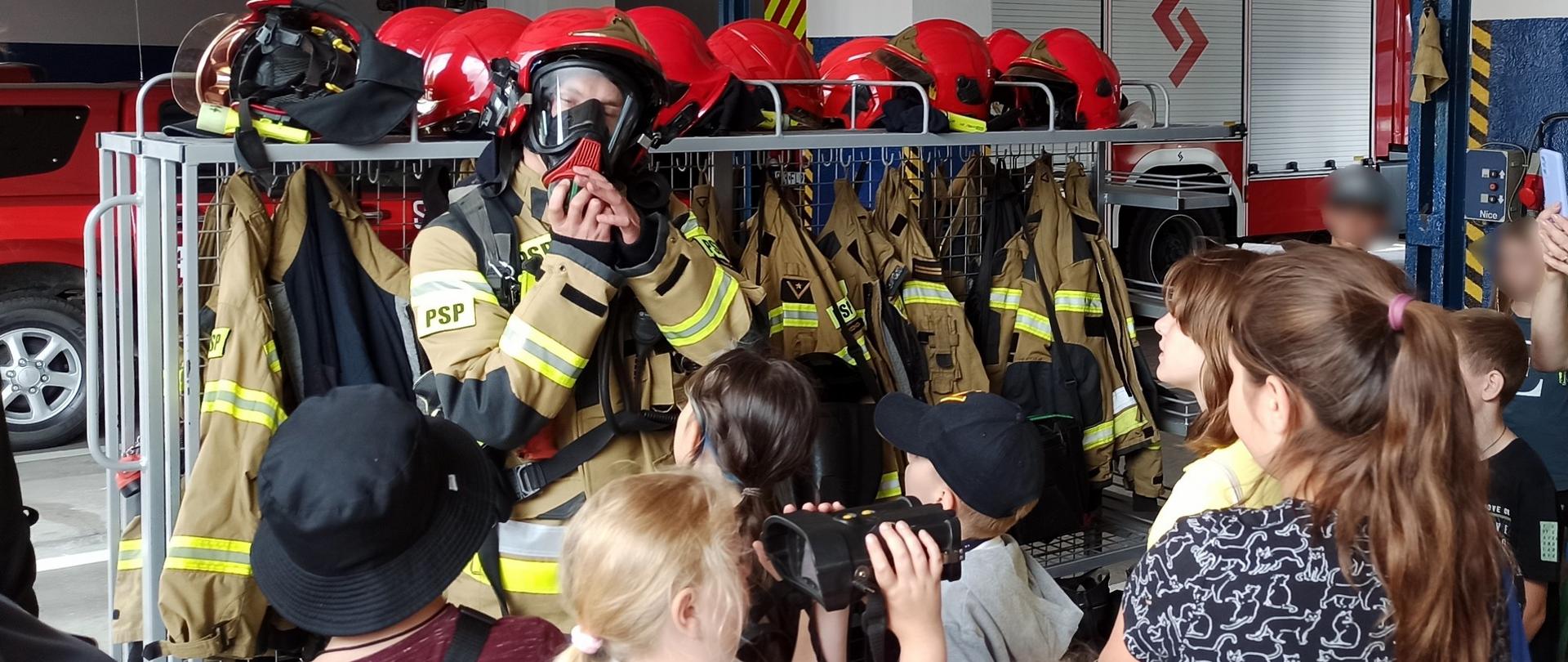 Zdjęcie przedstawia strażaka JRG Brodnica prezentującego obrania oraz sprzęt zabezpieczający podczas działań ratowniczo-gaśniczych. Przed strażakiem stoją zgromadzone dzieci.