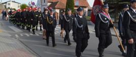 Dzień Strażaka w Sobiałkowie. Ulicą wioski na plac koło remizy maszerują z kościoła uczestnicy Mszy św. : poczet sztandarowy, Zarząd OSP, członkowie MDP oraz goście. W tle zabudowania.