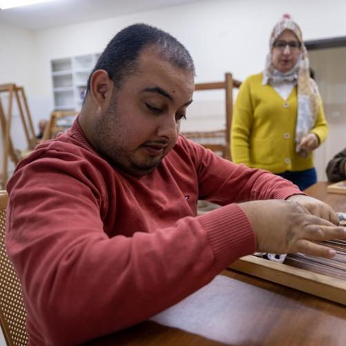 Na pierwszym planie mężczyzna w czerwonej buzie siedzi przy stole i tka na małym krośnie. W tle drugi mężczyzna robi to samo. Kobieta stoi i przygląda się.