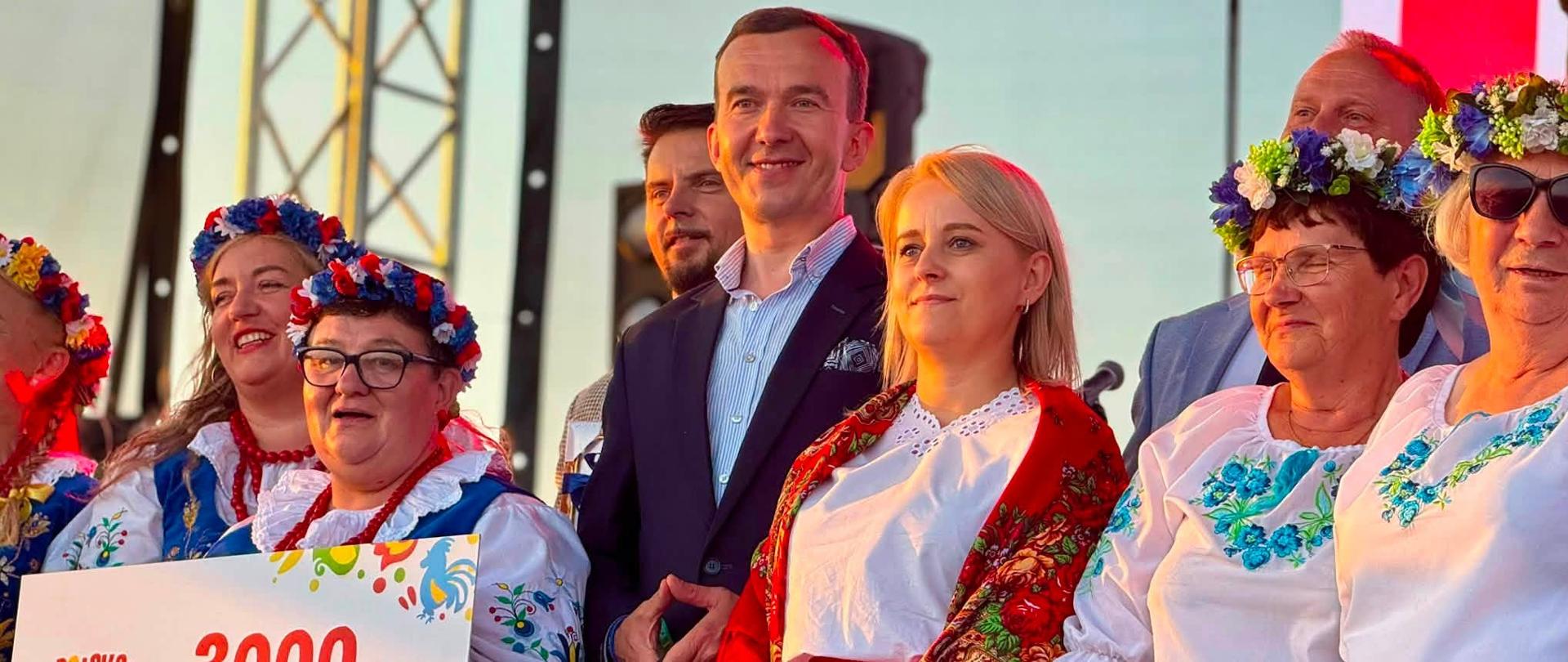 wicewojewoda stoi na scenie z kobietami w ludowych strojach trzymające czek pieniężny oraz inne nagrody 