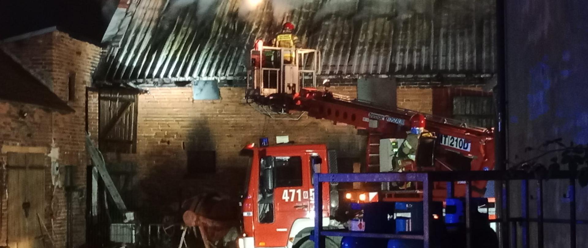 Pożar budynku gospodarczego, z dachu wydobywa się dym, przed budynkiem znajduje się strażacki podnośnik. W koszu podnośnika znajduje się strażak.