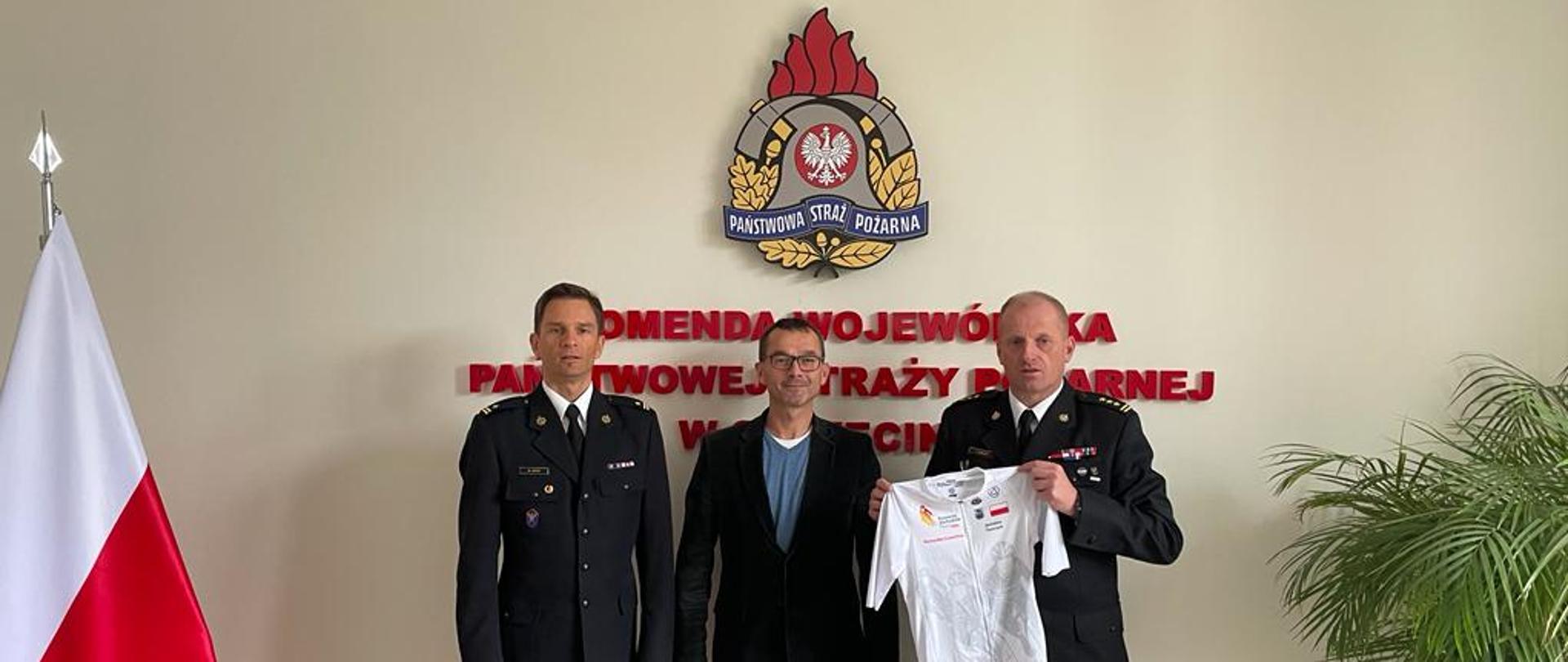 Na zdjęciu trzej mężczyźni na tle logo Państwowej Straży Pożarnej i Flagi Polski. Jeden z nich prezentuje koszulkę kolarską