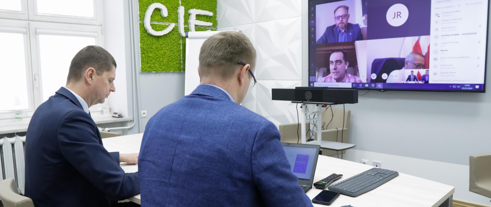 Dwóch mężczyzn w garniturach siedzi przy białym stole. W tle ekran z wideokonferencją.