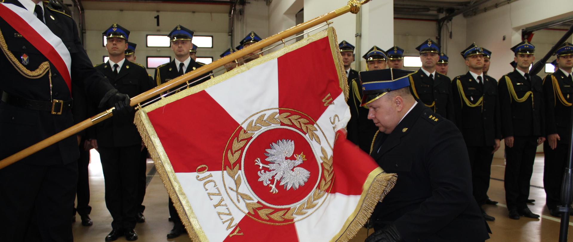 Powołany na stanowisko komendant w pozycji klęczącej całuje płat sztandaru. W tle pododdział strażaków KM PSP w Łomży.