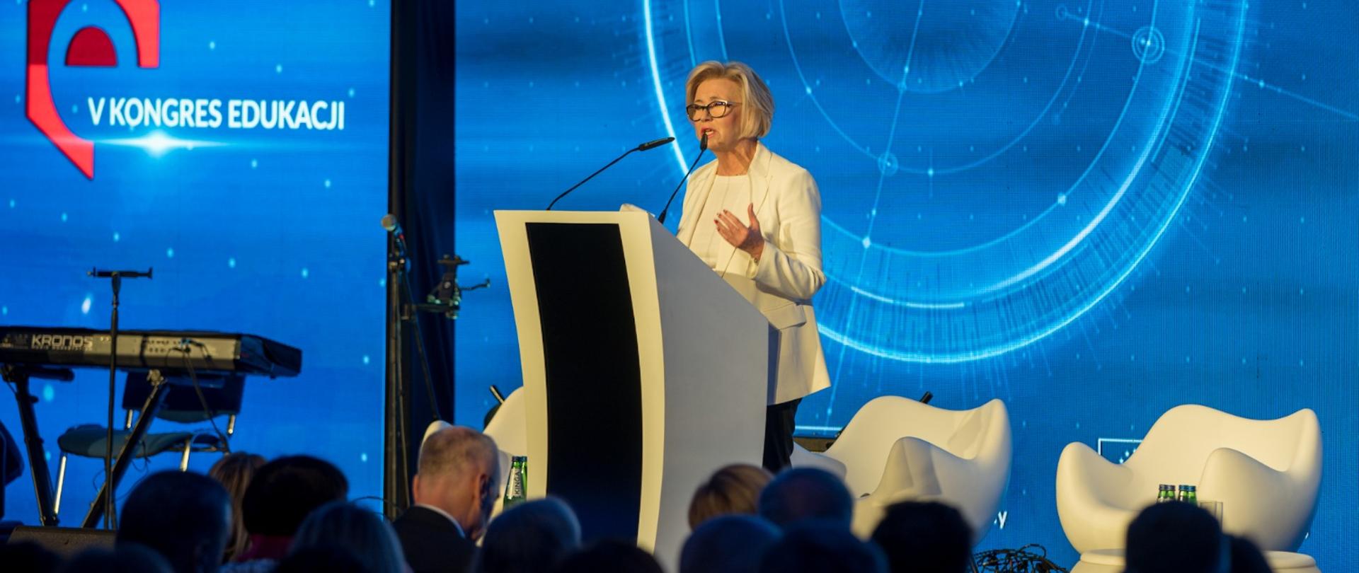 Wiceminister Machałek stoi przy mównicy, przed nią wypełniona ludźmi ciemna sala a za nią na ścianie napis V Kongres Edukacji.