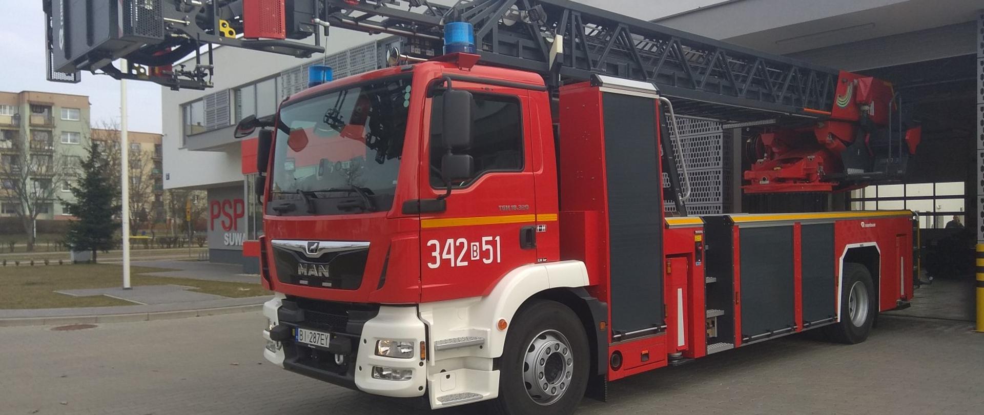 Samochód pożarniczy typu ciężkiego z koszem drabinowym o wysięgu 40 metrów. Pojazd koloru czerwony z białym zderzakiem. W tle budynek komendy i garaże.