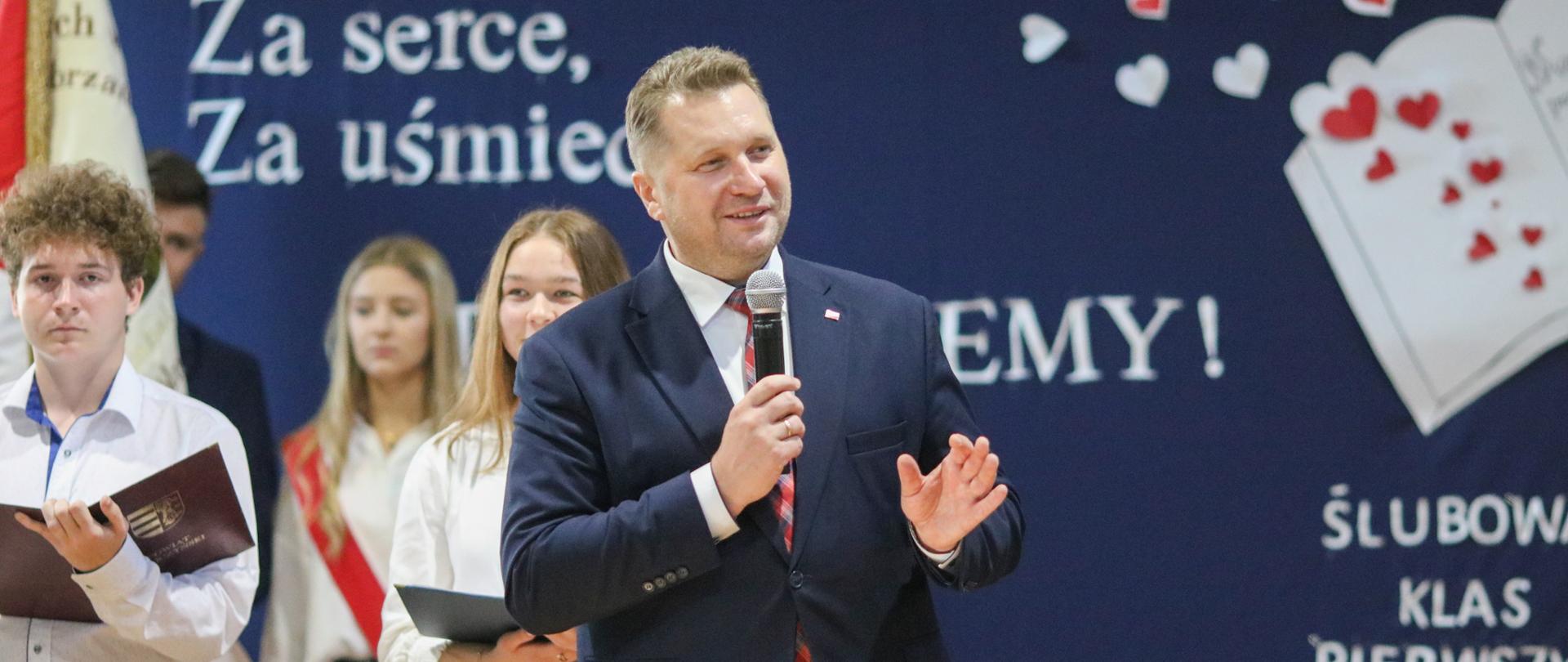 Minister Przemysław Czarnek stoi z mikrofonem i przemawia. Za nim dwójka młodzieży i poczet sztandarowy.