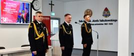 Pomorski komendant wojewódzki Państwowej Straży Pożarnej stoi w asyście swojego zastępcy oraz innego strażaka. Przed nimi stoi mikrofon. funkcjonariusze ubrani w mundury wyjściowe ze sznurem. 