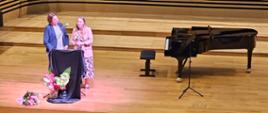 Dwie kobiety stoją na estradzie sali koncertowej przy mównicy i przemawiają do mikrofonu, za nimi po prawej stronie stoi fortepian.