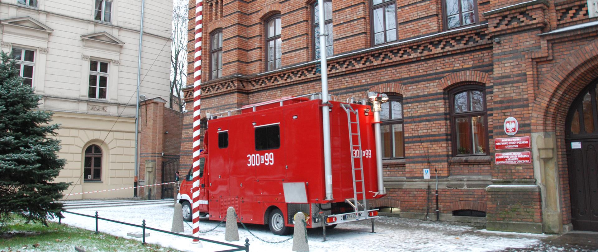 
Zdjęcie przedstawia pojazd dowodzenia i łączności koloru czerwonego, z rozstawionym masztem antenowym, przed budynkiem KM PSP w Krakowie.