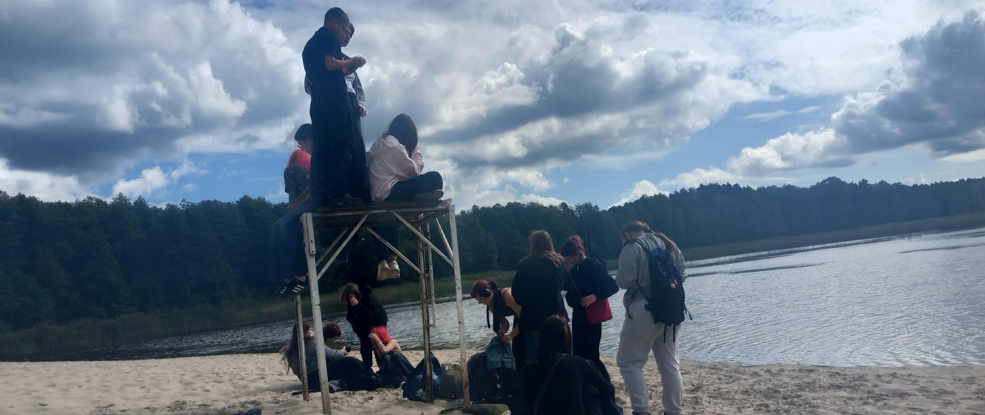 na zdjęciu jezioro otoczone lasem , na piaszczystej plaż grupa młodzieży pozuje do zdjęcia
