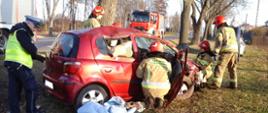 Uszkodzony samochód osobowy Toyota Yaris na poboczu drogi. Przy samochodzie pracujący strażacy. W tle stojący na jezdni samochód ratowniczo-gaśniczy.