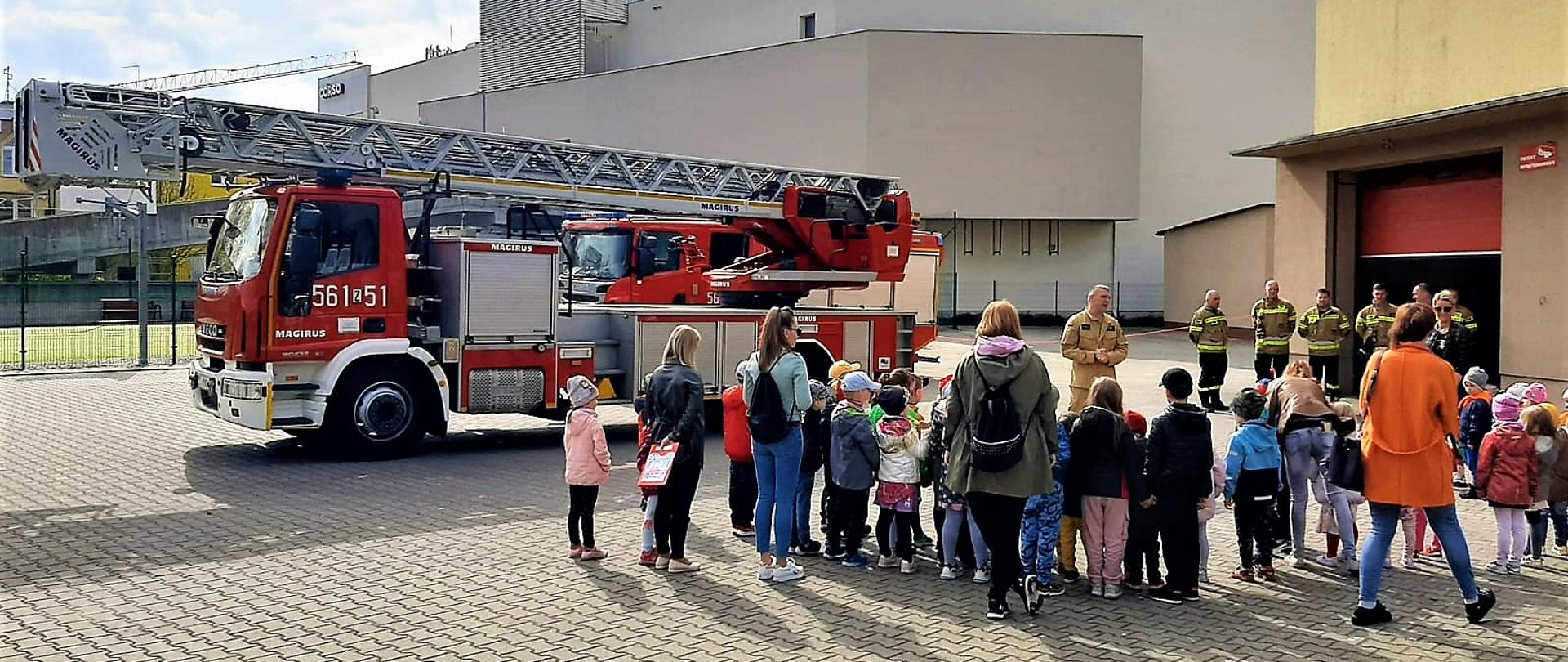 Strażacy w mundurach bojowych opowiadają grupie przedszkolaków i ich opiekunom o strażackiej służbie. W tle samochód pożarniczy drabina mechaniczna oraz budynek strażnicy.
