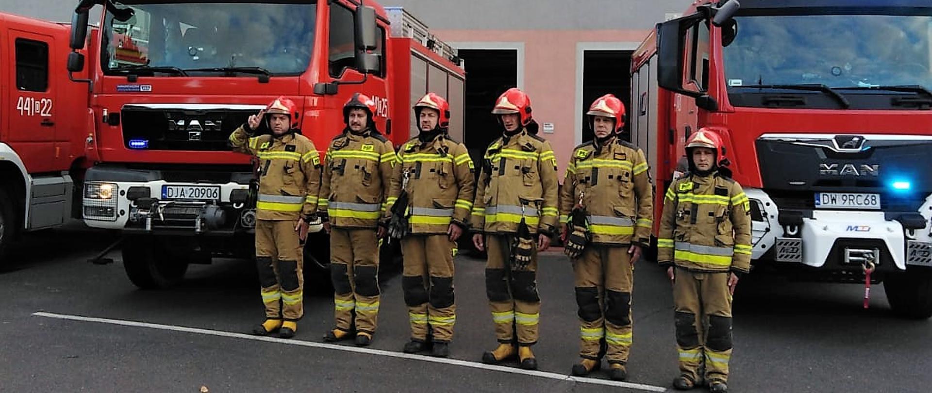 Obraz przedstawia strażaków jaworskiej komendy podczas minuty ciszy dla zmarłego strażaka. Strażacy w umundurowaniu specjalnym. W tle samochody pożarnicze i budynek komendy.