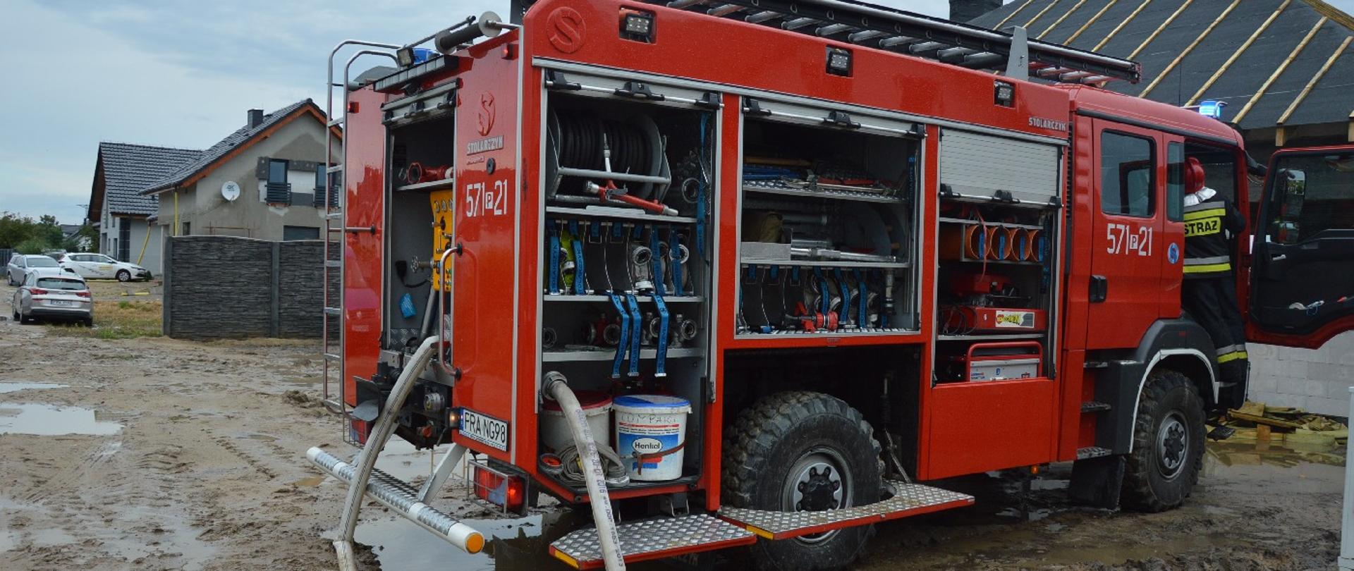 Wóz strażacki z otwartymi skrytkami. Podłączone są do niego 2 węże. W skrytkach widoczny sprzęt. Na drzwiach kabiny załogi numer 571P21. Drzwi do miejsca dowódcy otwarte. Na schodkach stoi odwrócony strażak. Wóz stoi na terenie błotnistym. Przed nim widoczny dach pokryty papą. Z lewej strony inne budynki mieszkalne oraz 3 samochody osobowe. 