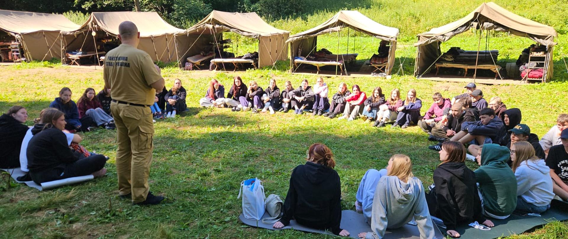 Zdjęcie przedstawia uczestników obozu harcerskiego pod namiotami oraz kadrę opiekunów podczas pogadanki edukacyjnej dla dzieci, młodzieży i osób dorosłych prowadzonej przez funkcjonariusza Komendy Powiatowej Państwowej Straży Pożarnej w Gołdapi