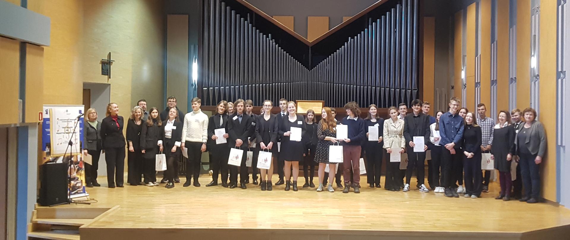Na scenie sali koncertowej wszyscy uczestnicy konkursu wraz z organizatorami oraz jurorami. Na pierwszym planie fotele. Na scenie uczestnicy trzymają w dłoniach dyplomy a laureaci dodatkowo torby z nagrodami. Po lewej stronie mikrofon oraz kolumna nagłośniająca oraz plakat informujący o konkursie. W tle organy