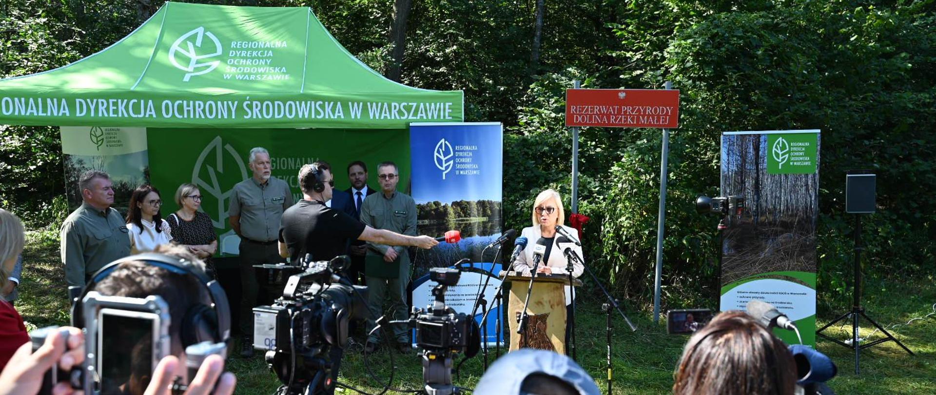 Ministra klimatu i środowiska Paulina Hennig-Kloska podczas konferencji prasowej dotyczącej ustanowienia 200. nowego rezerwatu przyrody od 2024 roku. Na zdjęciu ministra stoi przy mównicy i mówi do mikrofonów. Konferencja odbywa się na zewnątrz, a na pierwszym planie zdjęcia widać dziennikarzy i kamery.