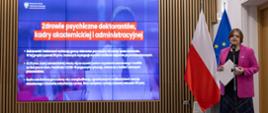 Na tle ściany z pionowych drewnianych listewek z wbudowanym wielkim ekranem z napisem na niebieskim tle "Zdrowie psychiczne doktorantów, kadry akademickiej i administracyjnej" stoi wiceminister Zioło-Pużuk w różowej marynarce i mówi do mikrofonu na stojaku.