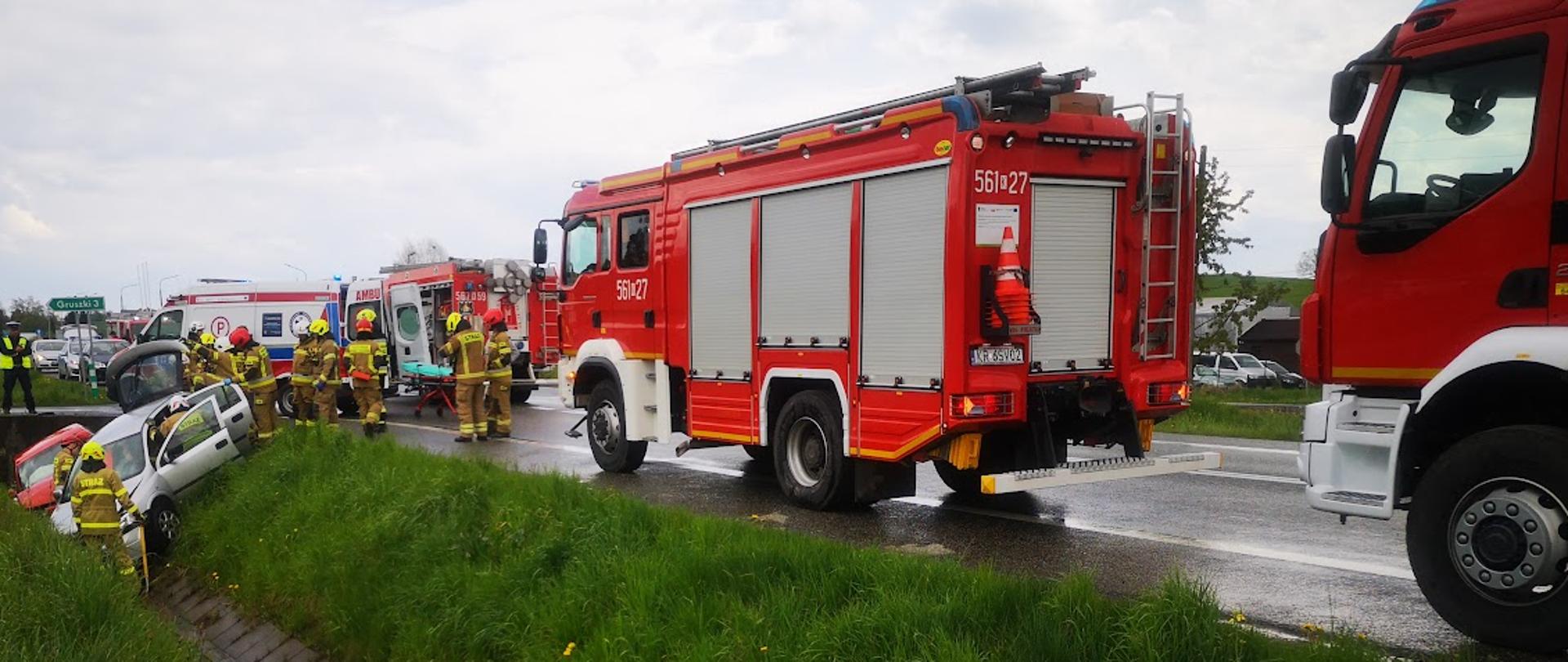 Na zdjęciu dwa samochody osobowe w rowie, na drodze samochody pożarnicze. Przy samochodach osobowych strażacy prowadzący działania ratownicze