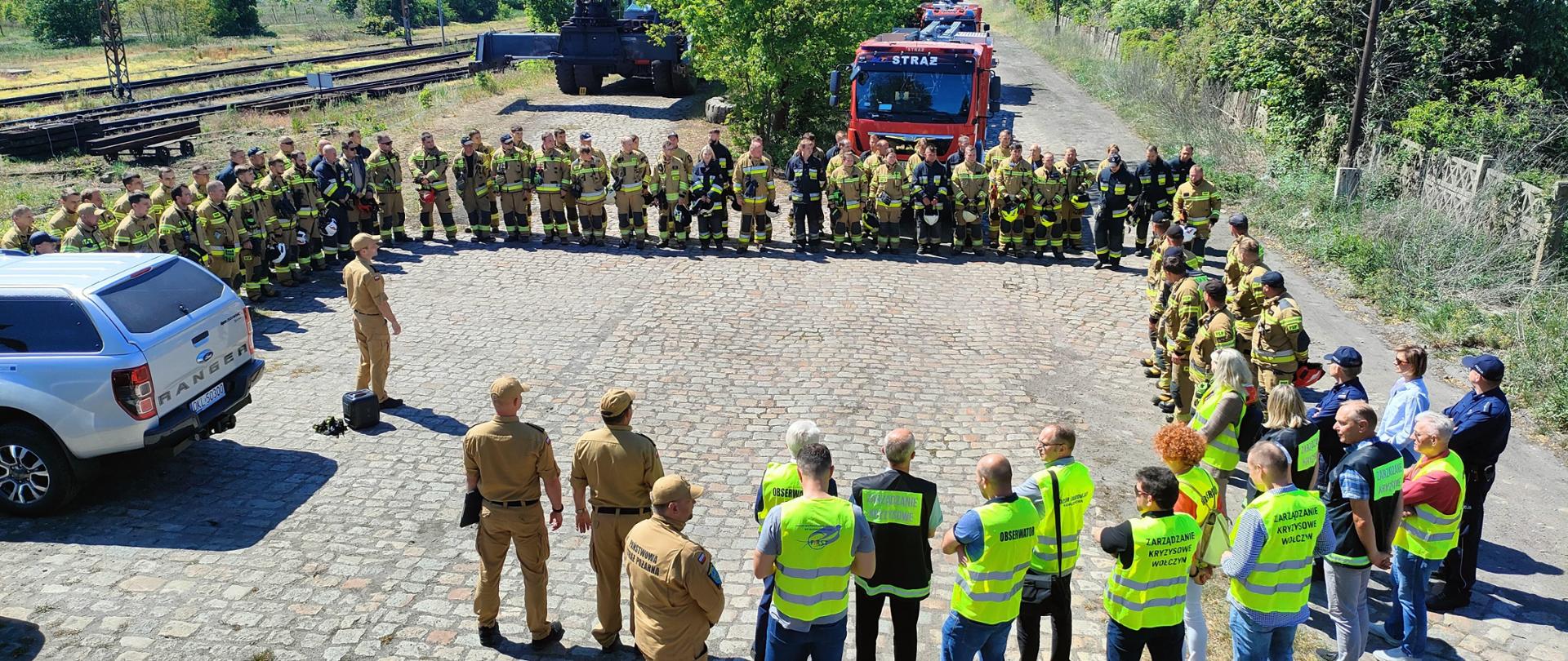 Na zdjęciu znajduje się grupa ludzi uczestniczących w odprawie przed ćwiczeniami na świeżym powietrzu. Widoczne są dwie grupy osób: jedna składająca się z strażaków ubranych w pełne mundury ochronne, stojąca w półkolu, oraz druga grupa osób w kamizelkach odblaskowych z napisami takimi jak "Zarządzanie Kryzysowe" i "Obserwator", która stoi naprzeciwko nich. Na pierwszym planie widocznych jest kilku mężczyzn w beżowych mundurach. W tle widać zaparkowane wozy strażackie, teren porośnięty roślinnością oraz tory kolejowe. Pogoda jest słoneczna, a niebo jasne.