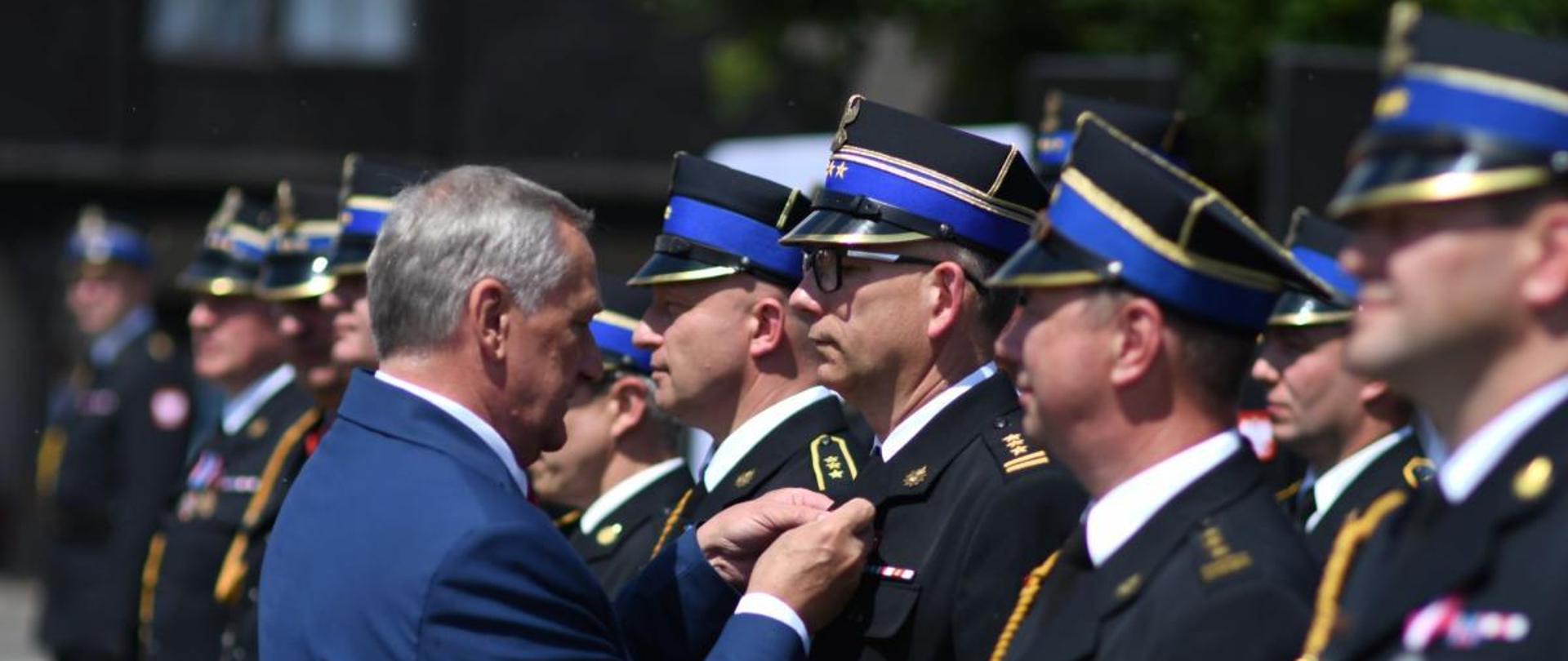 W szeregu stoi grupa mężczyzn ubranych w wyjściowe mundury Państwowej Straży Pożarnej, przed nimi mężczyzna w stroju cywilnym przypina medal jednemu ze strażaków