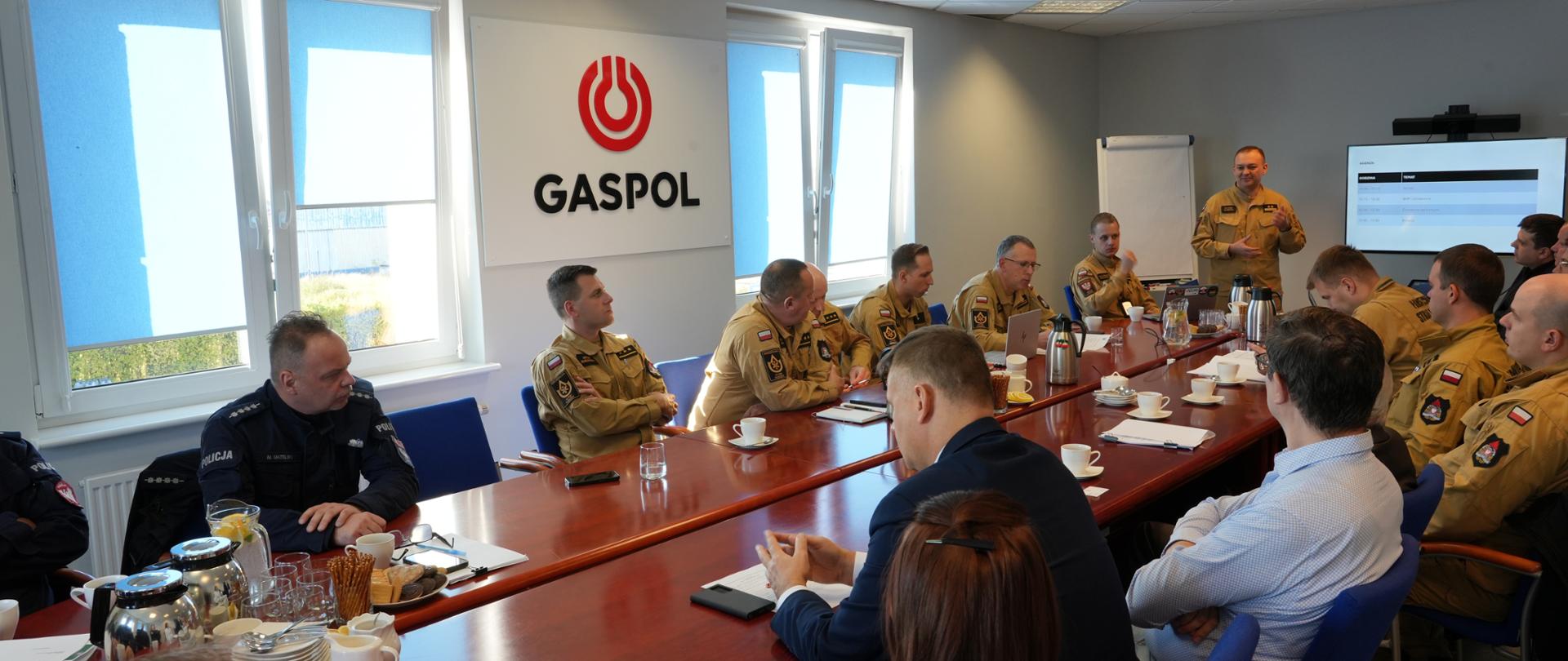 Szeroki kadr sali konferencyjnej z widocznym na ścianie logo GASPOL. Przy stole obradują strażacy, policjant oraz cywile, a na końcu stołu stoi przemawiający strażak.