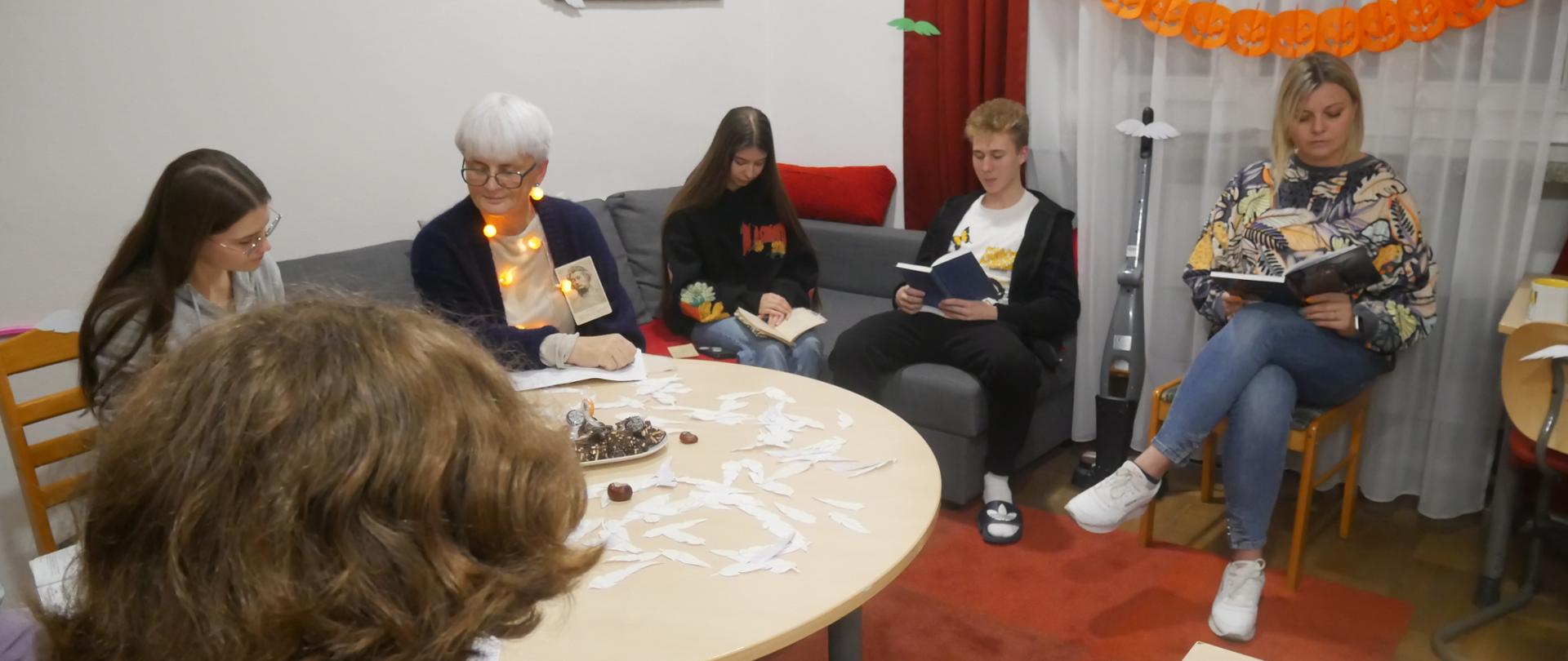 W spotkaniu uczestniczy również p. Dyrektor – pierwsza od prawej strony. Obok p. Dyrektor siedzi Janek, Agnieszka i wspomniane wcześniej osoby. Wszyscy wpatrzeni są w książki. W oknie na firanie wisi dyniowy łańcuch, a po pokoju fruwają duchy. 