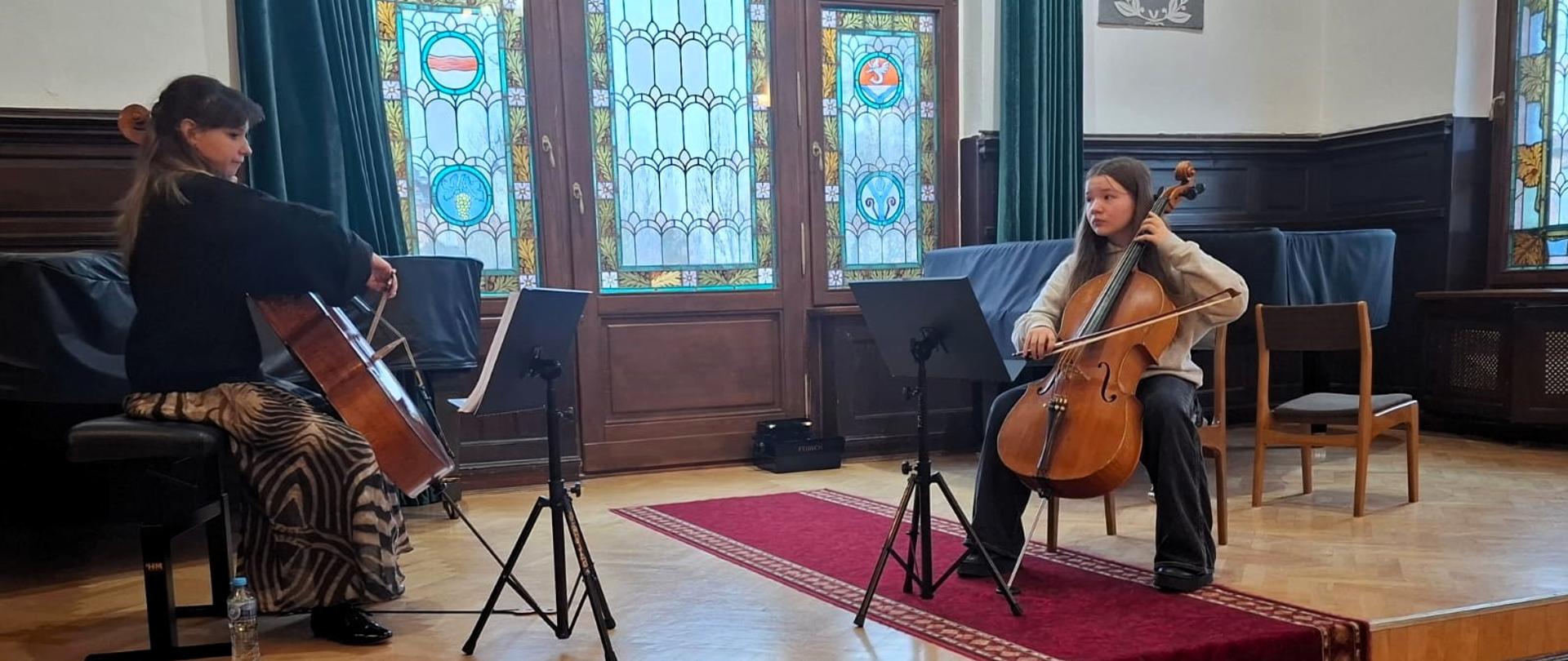 Na scenie auli szkolnej dwie osoby - młoda kobieta oraz uczennica wieku około 12 lay ćwiczą grę na wiolonczeli. Obie siedzą na krzesłach i grają na instrumentach. Przed nimi stoją pulpity z nutami. w tle można zaobserwować dwa fortepiany, duże okno z kolorowym witrażem ora zielone zasłony. Na podłodze leży czerwony dywan.