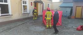 Tragiczny bilans pożaru. Posesja w Rawiczu przy ul. Piłsudskiego. Na podwórzu, przed kamienicą ustawiony jest parawan ochronny koloru czerwonego. Drzwi wejściowe oraz dwa okna kamienicy są otwarte. Na podwórku dwóch strażaków i mieszkaniec kamienicy.