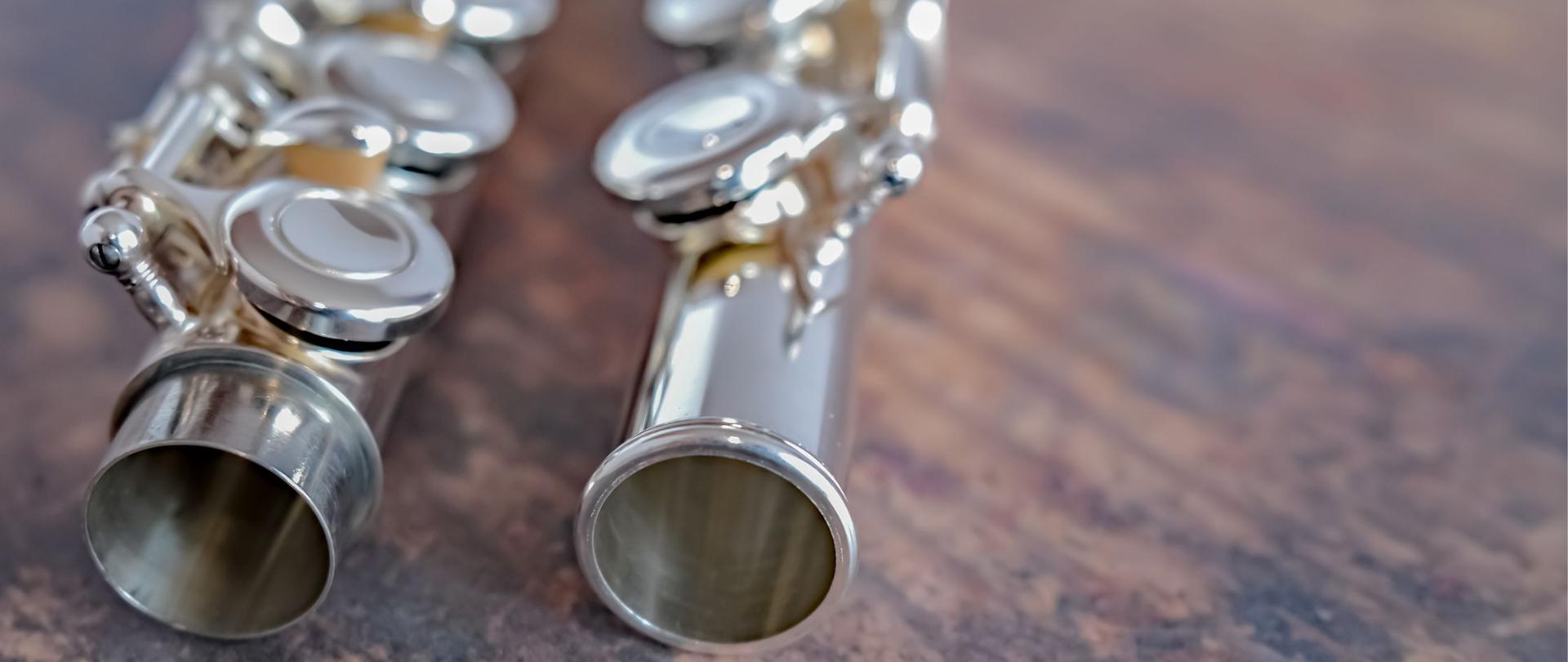 Zbliżenie na dwa metalowe flety poprzeczne leżące na drewnianej powierzchni, z widocznymi klapami i otwartymi końcówkami instrumentów.