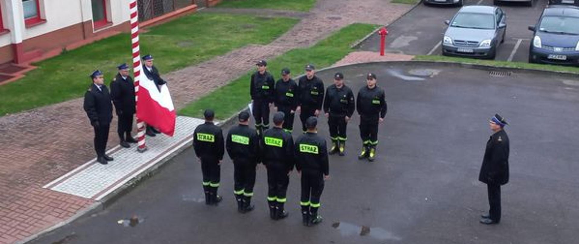 Uroczysta zmiana służby z okazji Narodowego Święta Niepodległości. Strażacy ze zmian służbowych stoją naprzeciwko siebie. Poczet flagowy wciąga flagę państwową na maszt. 