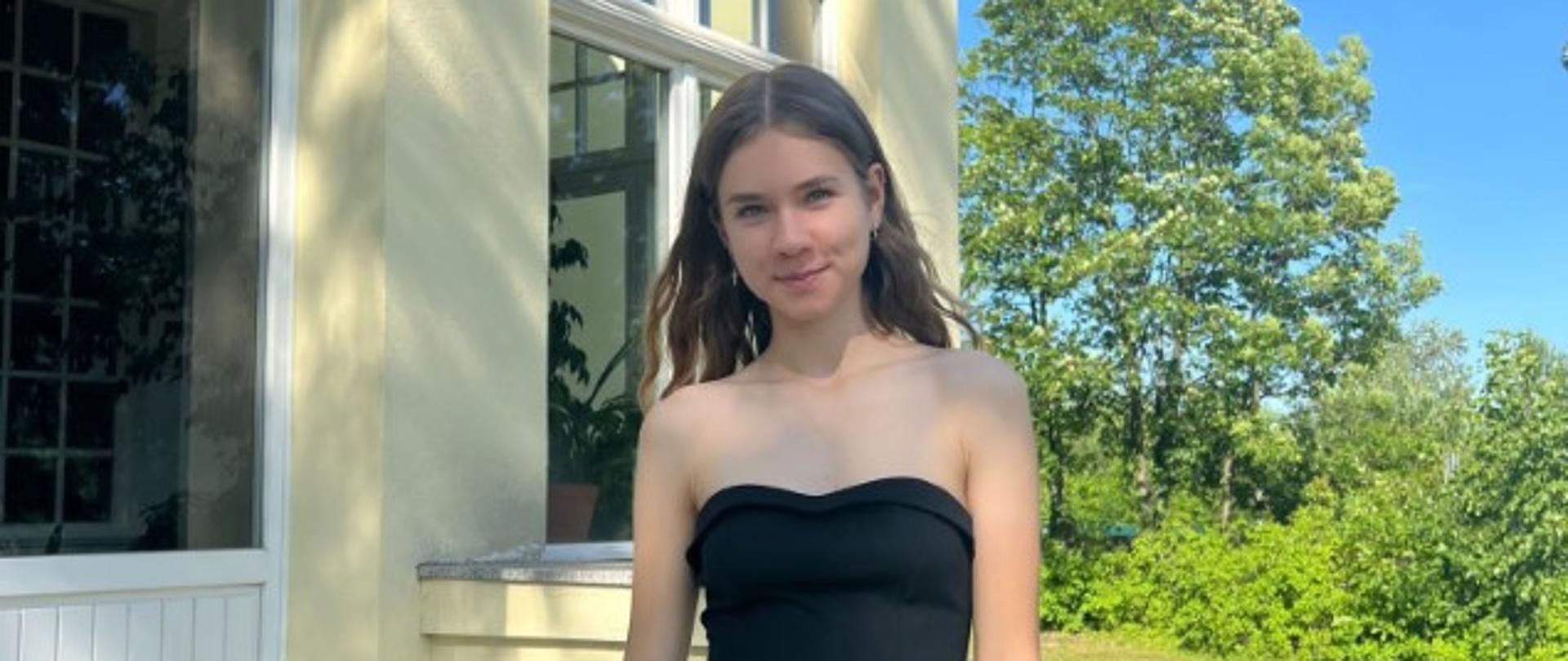 Zdjęcie kolorowe. Nastolatka w czarnym stroju stoi na schodach. W tle zielona roślinność.