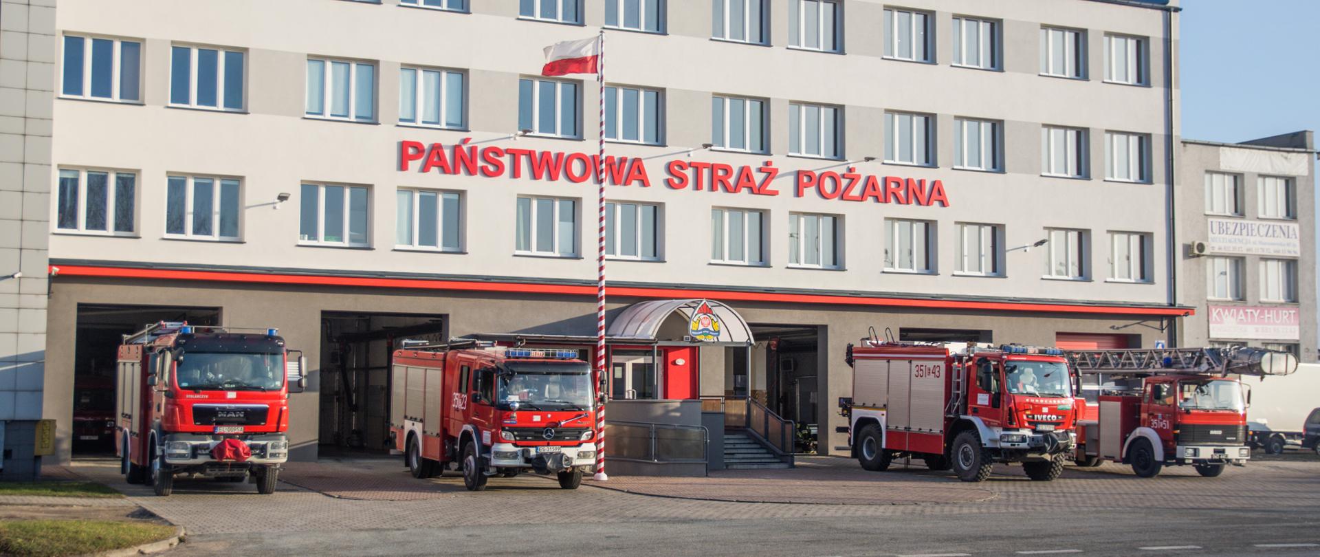 Zdjęcie przedstawia widok budynku komendy po remoncie przeprowadzonym w 2020 roku. Przed budynkiem zaparkowane są pojazdy pożarnicze jednostki ratowniczo-gaśniczej. Na maszcie podniesiona jest flaga państwowa.