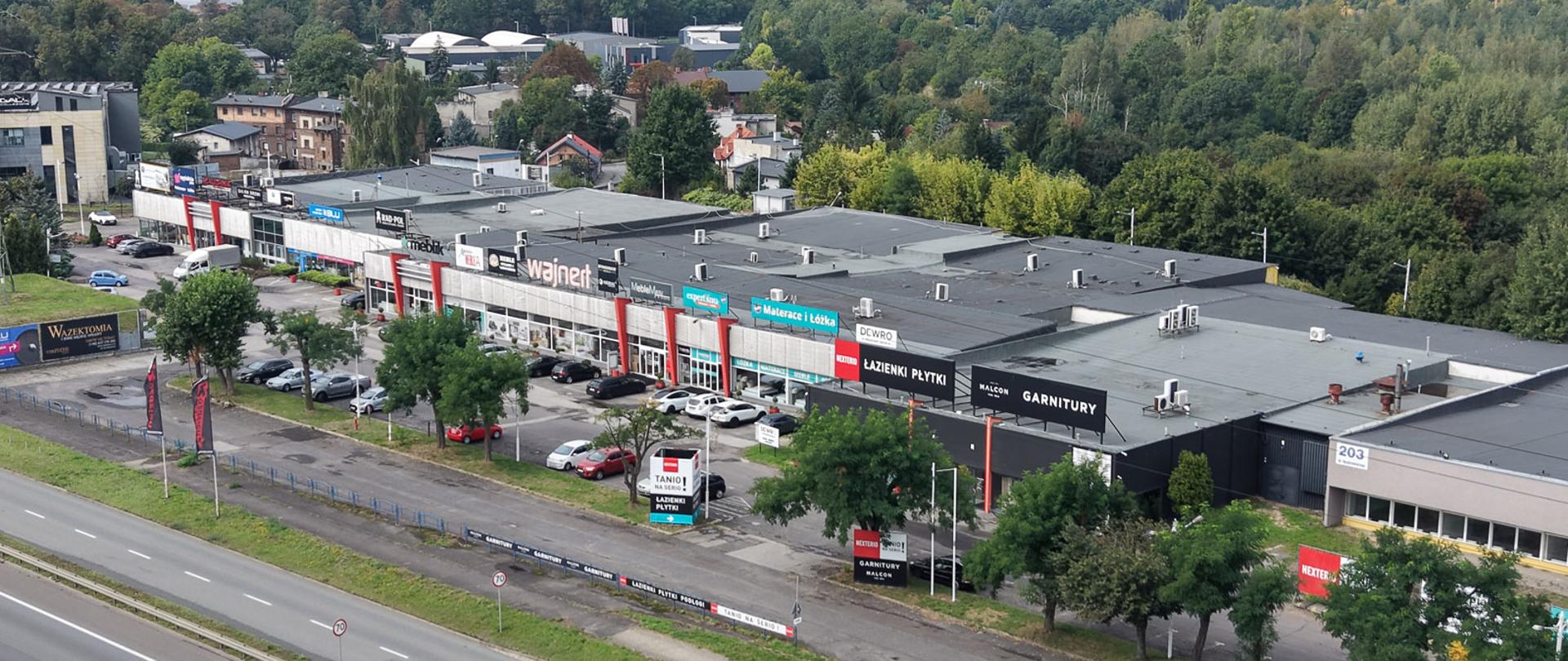 Dzień latem, zbliżenie z drona na parterowe zabudowania budynków handlowych w zwartej zabudowie, nad witrynami szyldy reklamowe, przed budynkami parking i samochody, a w kolejnej linii wielopasmowa droga.