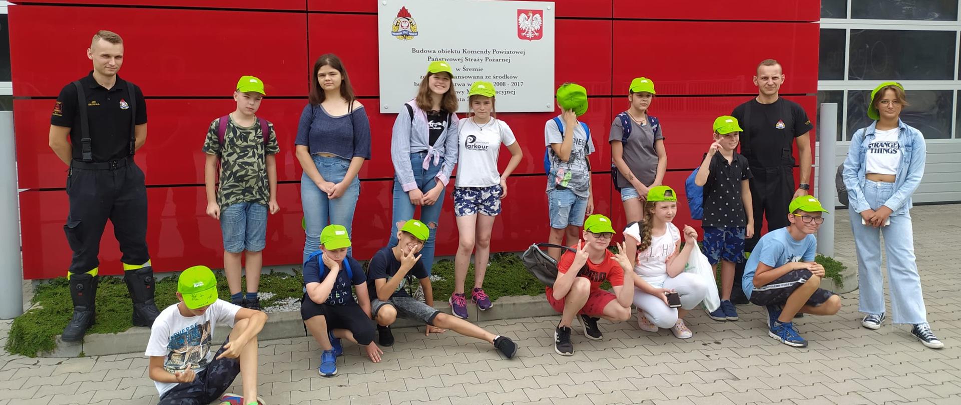 Zdjęcie na tle komendy powiatowej PSP w Śremie dzieci z półkolonii opiekunowie oraz dwóch strażaków a na ścianie budynku tablica informacyjna o budowie i finansowaniu 