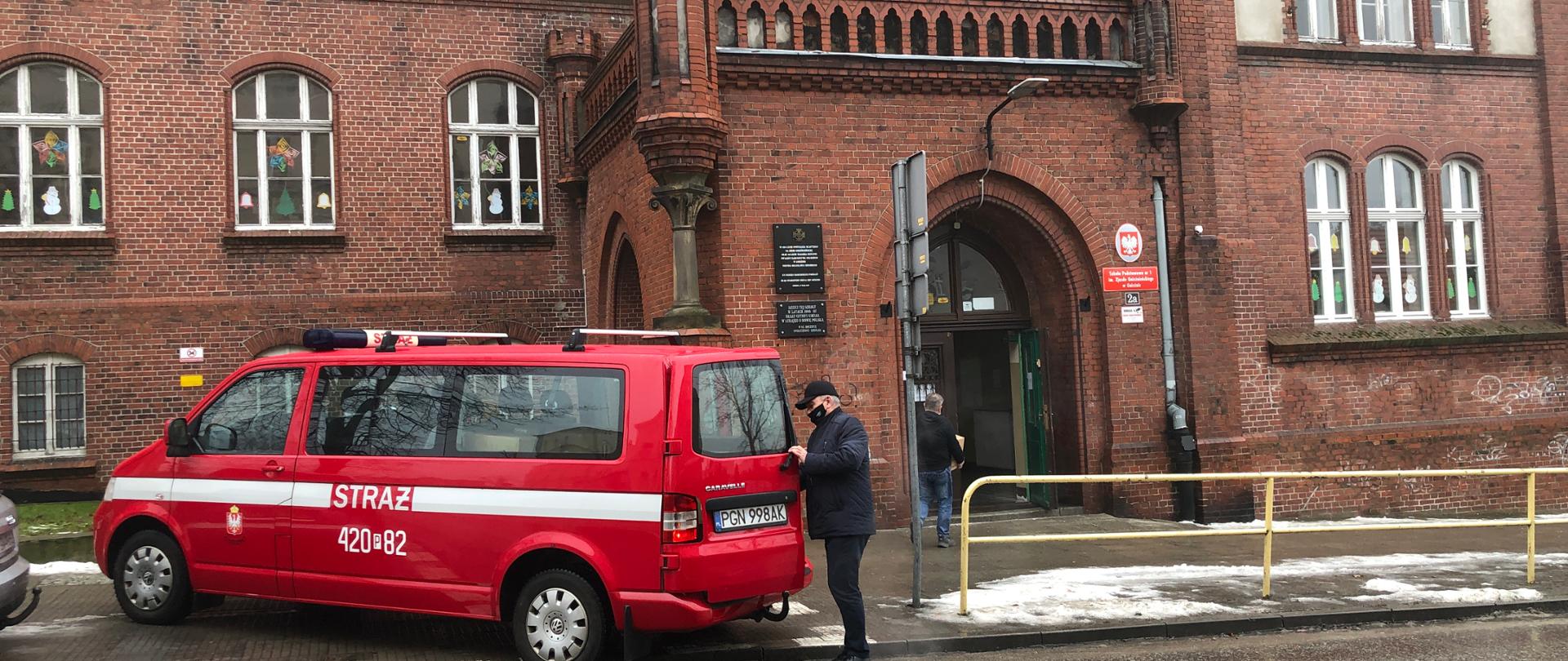 Samochód strażacki bus koloru czerwonego zaparkowany przed budynkiem szkoły. Przy tylnych drzwiach samochodu stoi mężczyzna, który przez lekko uchylone drzwi zagląda w część bagażową. Za busem widoczny budynek szkoły podstawowej. Drzwi główne do szkoły są otwarte a do wejścia idzie osoba, która niesie karton