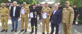 Zdjęcie przedstawia strażaków oraz osoby cywilne podczas wręczenia pucharów i dyplomów za zajęcie pierwszego miejsca. 