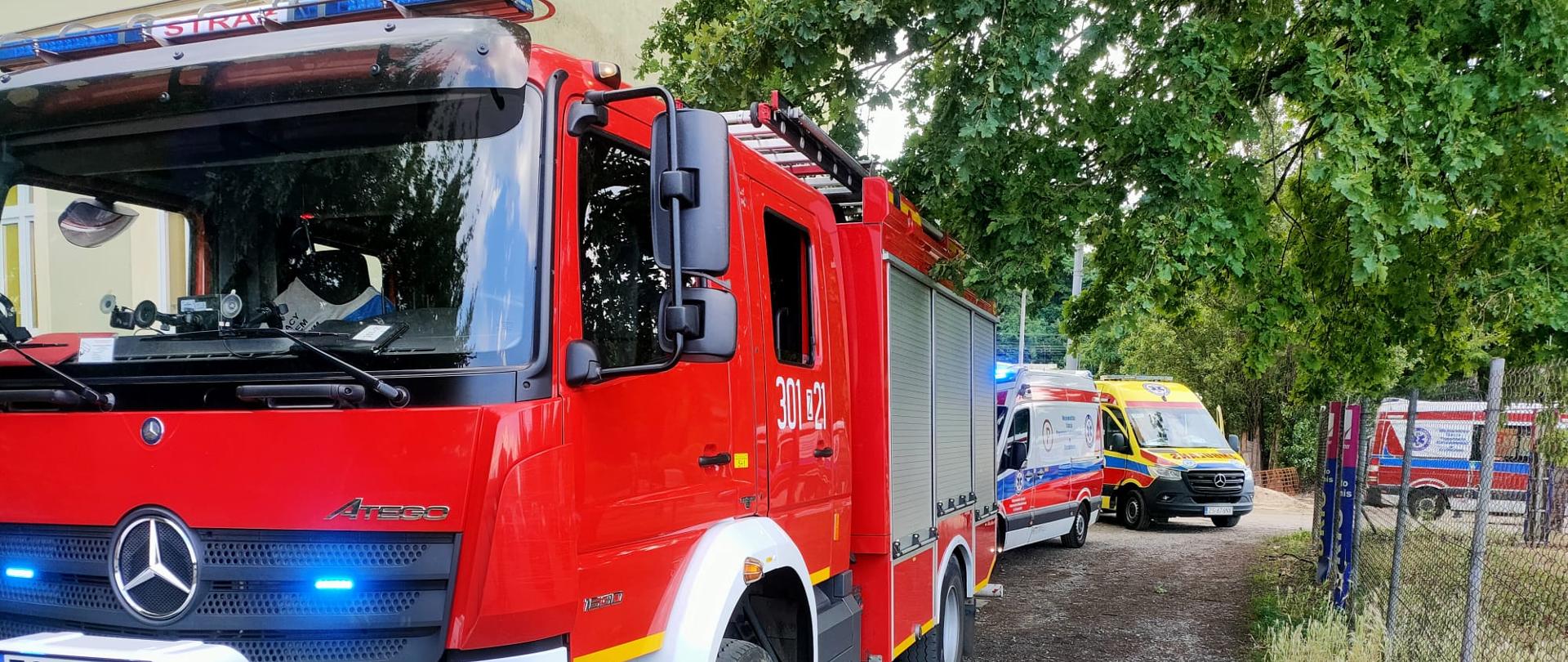 Samochód pożarniczy bez strażaków z włączanymi światłami błyskowymi. W tle stoją trzy karetki pogotowia ratunkowego.