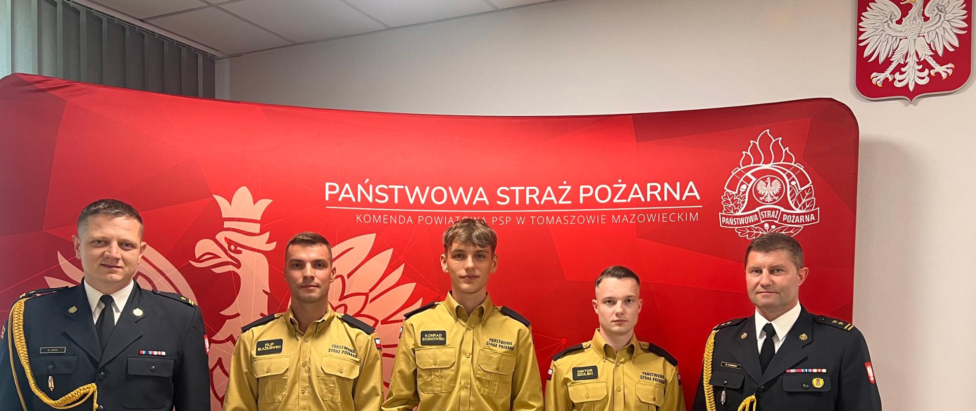 Strażacy w mundurach wyjściowych stoją na skrajnych częściach zdjęcia. Po środku trzech mężczyzn w piaskowych ubraniach trzymający teczkę. W tle czerwony parawan z orłem herbem Państwowej Straży Pożarnej oraz napisem Państwowa straż Pożarna. 