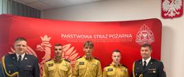 Strażacy w mundurach wyjściowych stoją na skrajnych częściach zdjęcia. Po środku trzech mężczyzn w piaskowych ubraniach trzymający teczkę. W tle czerwony parawan z orłem herbem Państwowej Straży Pożarnej oraz napisem Państwowa straż Pożarna. 