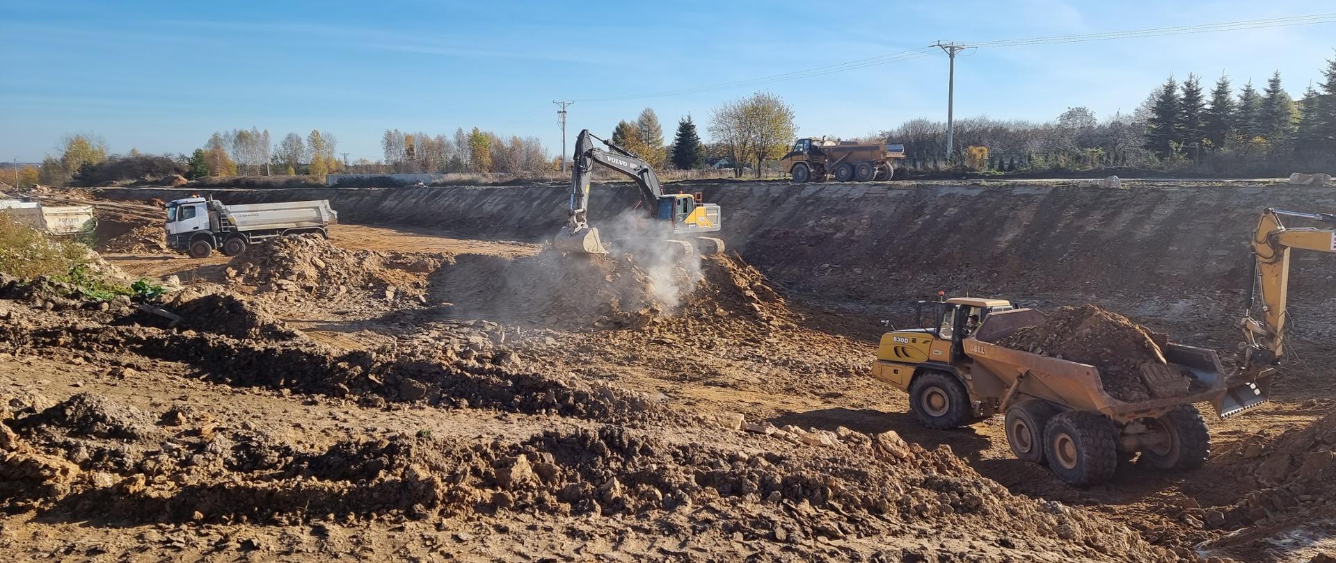 DK42 obwodnica Wąchocka - budowa, wykop z gruncie skalistym, dwie pracujące koparki, samochody ciężarowe wywożące grunt 
