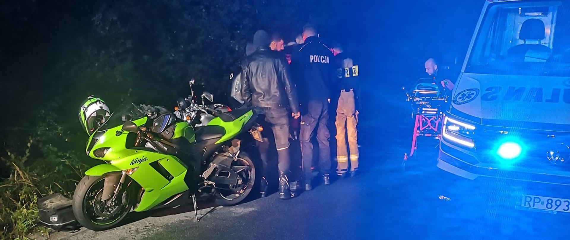 Na zdjęciu widać jeden motocykl przy którym stoi grupa osób w kółku oraz karetka na sygnałach. Zdjęcie wykonane jest w nocy.