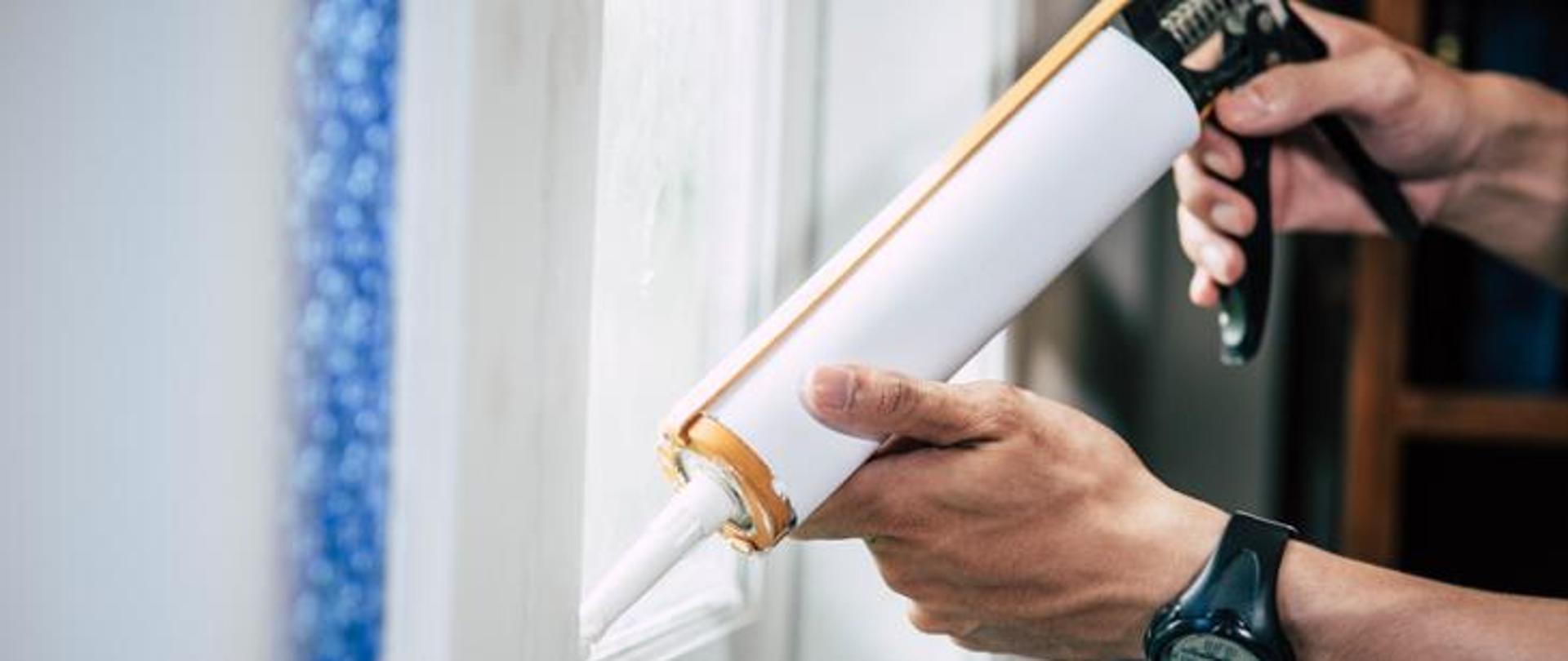 Na zdjęciu widać dłonie trzymające tubkę sylikonu. Osoba za pomocą tak zwanego pistoletu wyciska sylikon uszczelniając okno.