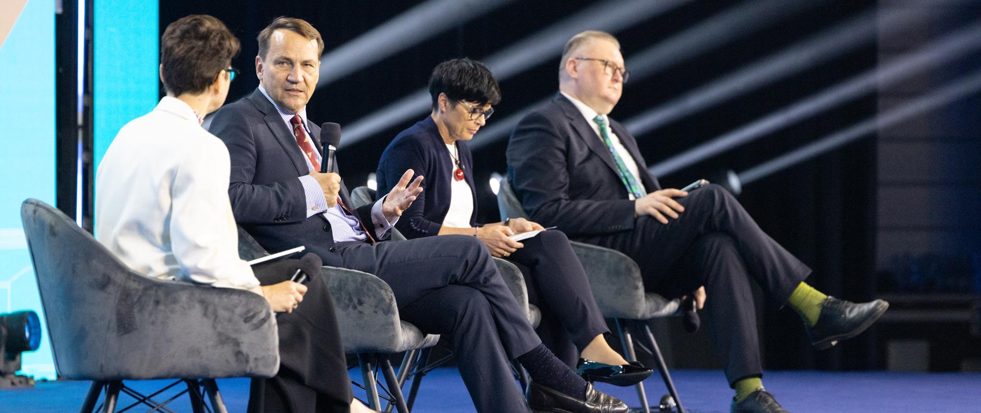 Cztery osoby uczestniczą w panelu dyskusyjnym na scenie konferencyjnej. Wicepremier Sikorski w granatowym garniturze, jasnoniebieskiej koszuli i czerwonym krawacie mówi do mikrofonu, gestykulując dłonią.