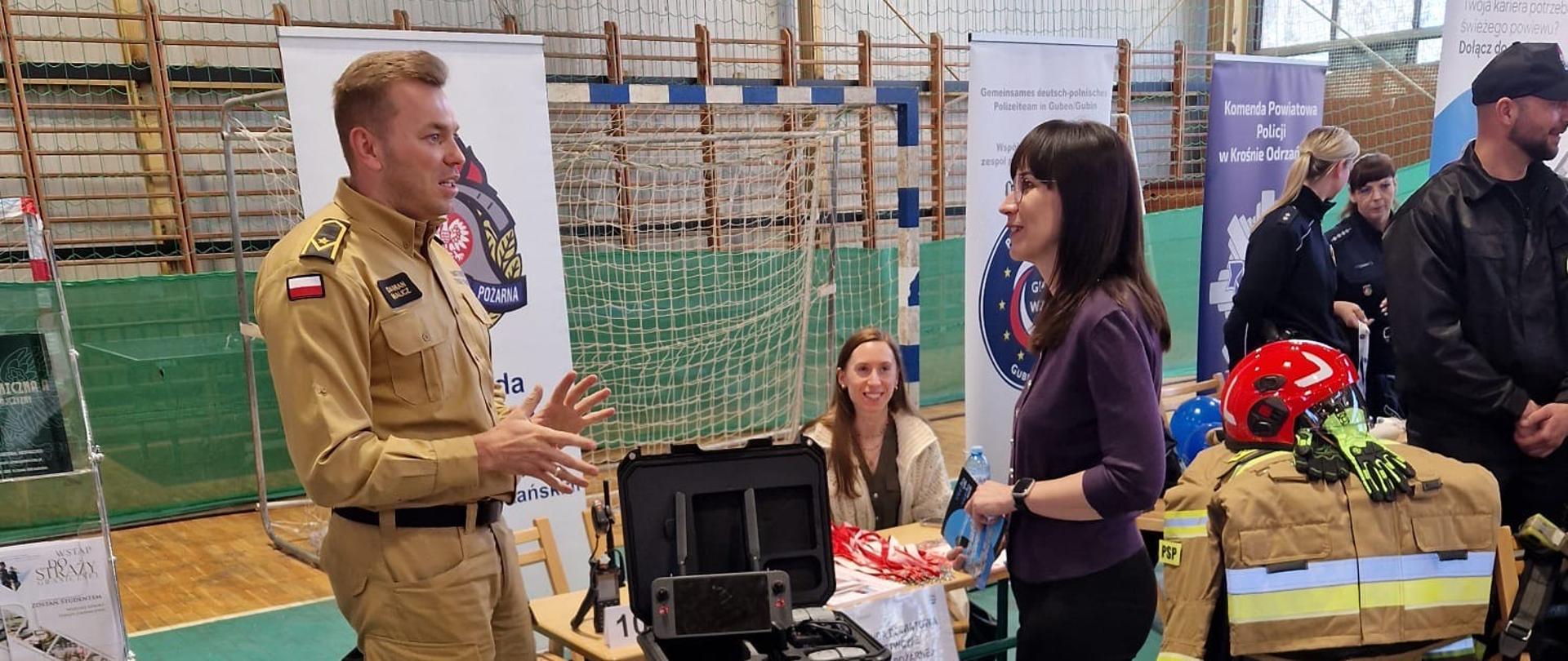 Strażak z Komendy Powiatowej Państwowej Straży Pożarnej w Krośnie Odrzańskim prezentuje sprzęt ratowniczy przy stanowisku podczas Targów Pracy. Na stole widoczny jest dron i walizka ze sprzętem, a na podłodze leży fantom do nauki udzielania pierwszej pomocy. W tle przedstawiciele innych służb i odwiedzający.