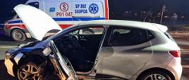 rozbity biały samochód z podniesioną maską oraz wyrwanymi drzwiami kierowcy za nim stoi karetka pogotowia